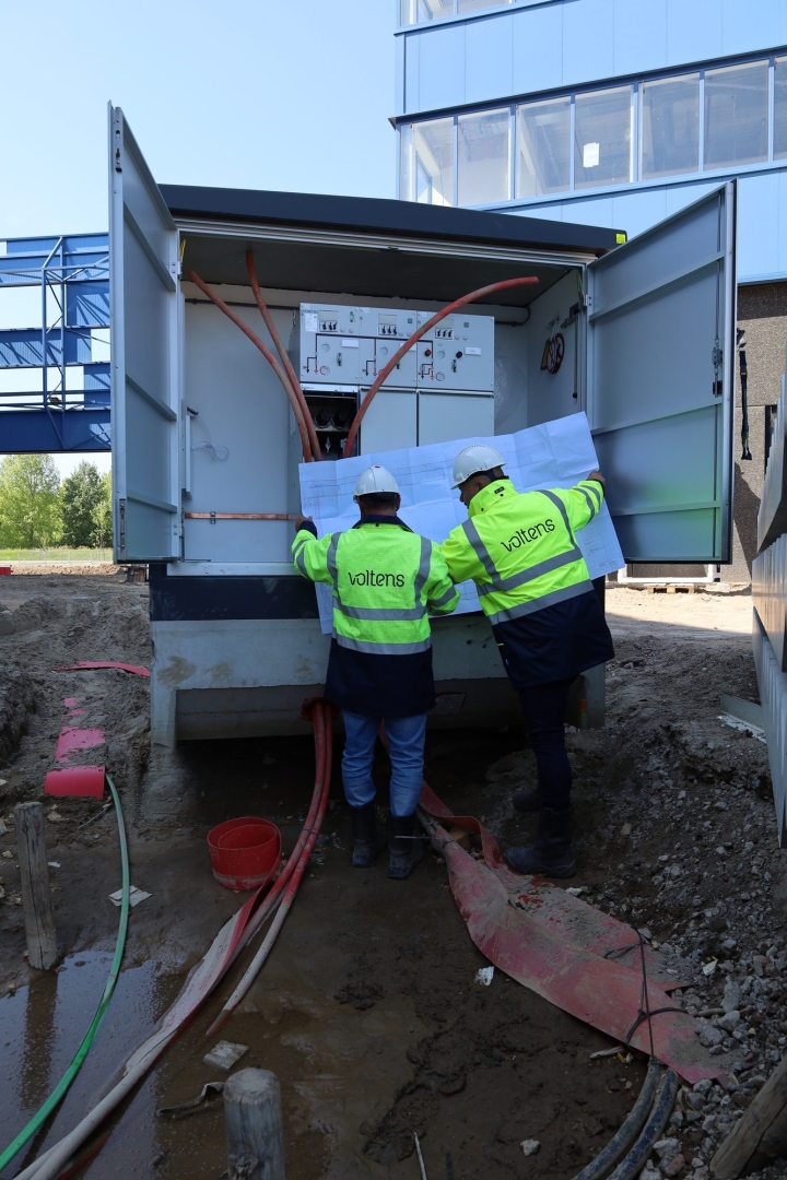 Twee monteurs in Voltens-werkkleding bestuderen een tekening voor een geopend compactstation